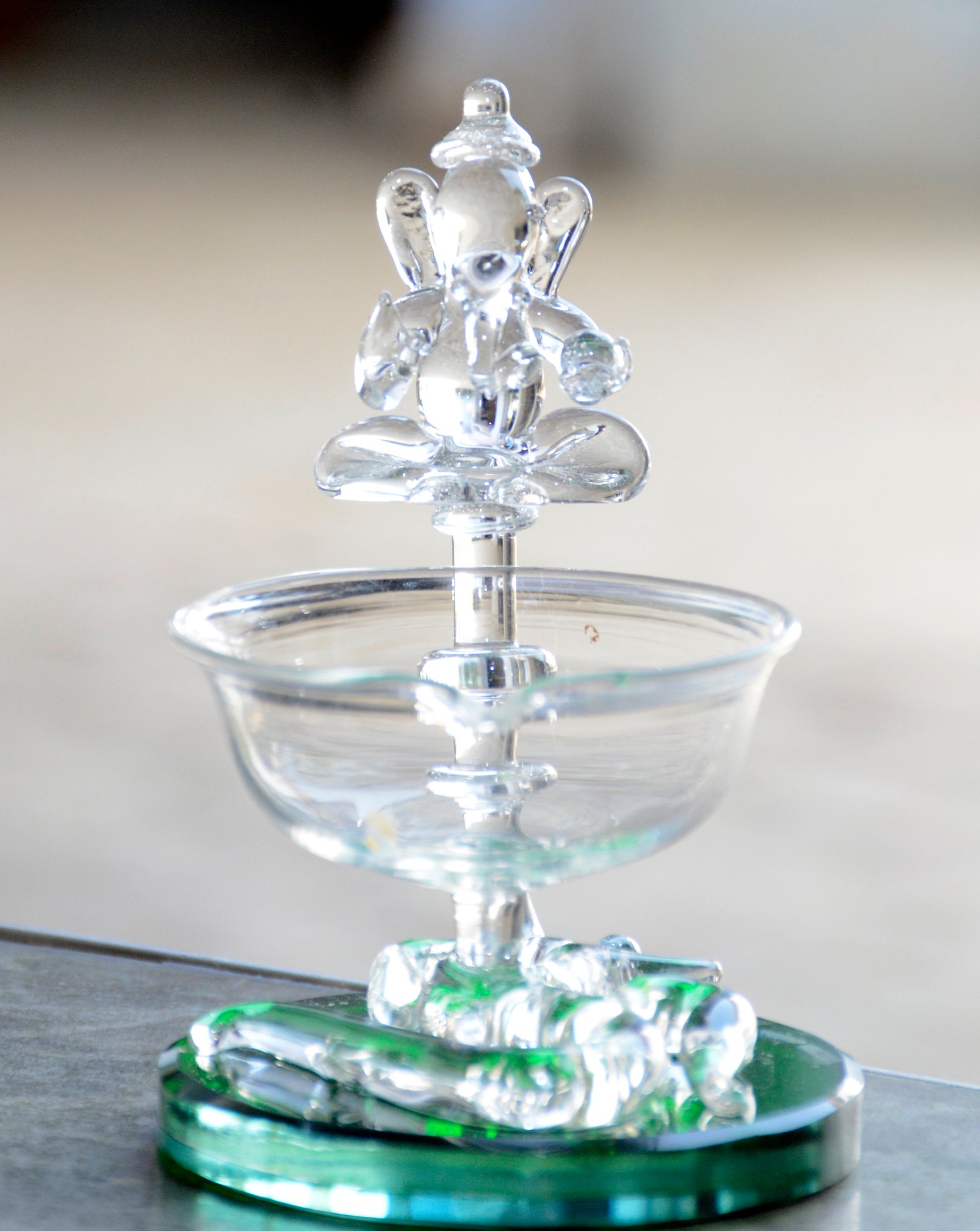 A small, decorative glass diya (oil lamp) featuring a representation of Lord Ganesha, designed for holding a flame.