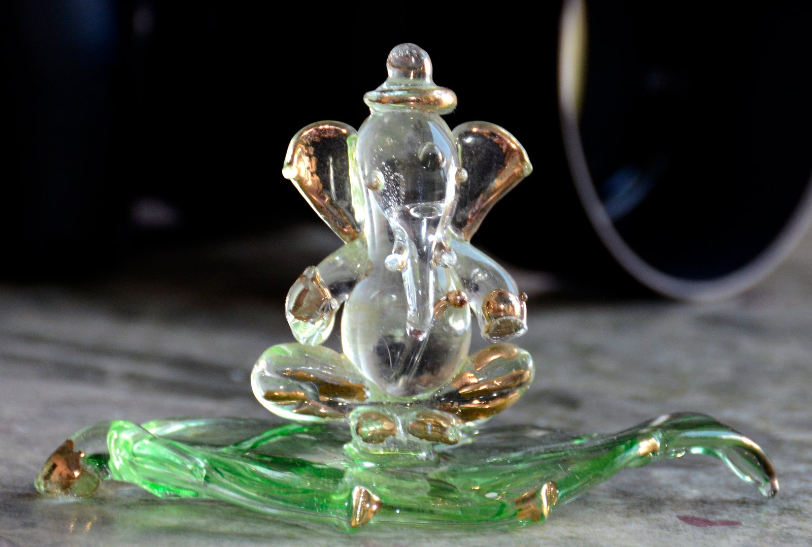 A small, translucent glass representation of Lord Ganesha, featuring subtle gold accents and sitting atop a decorative green glass base.