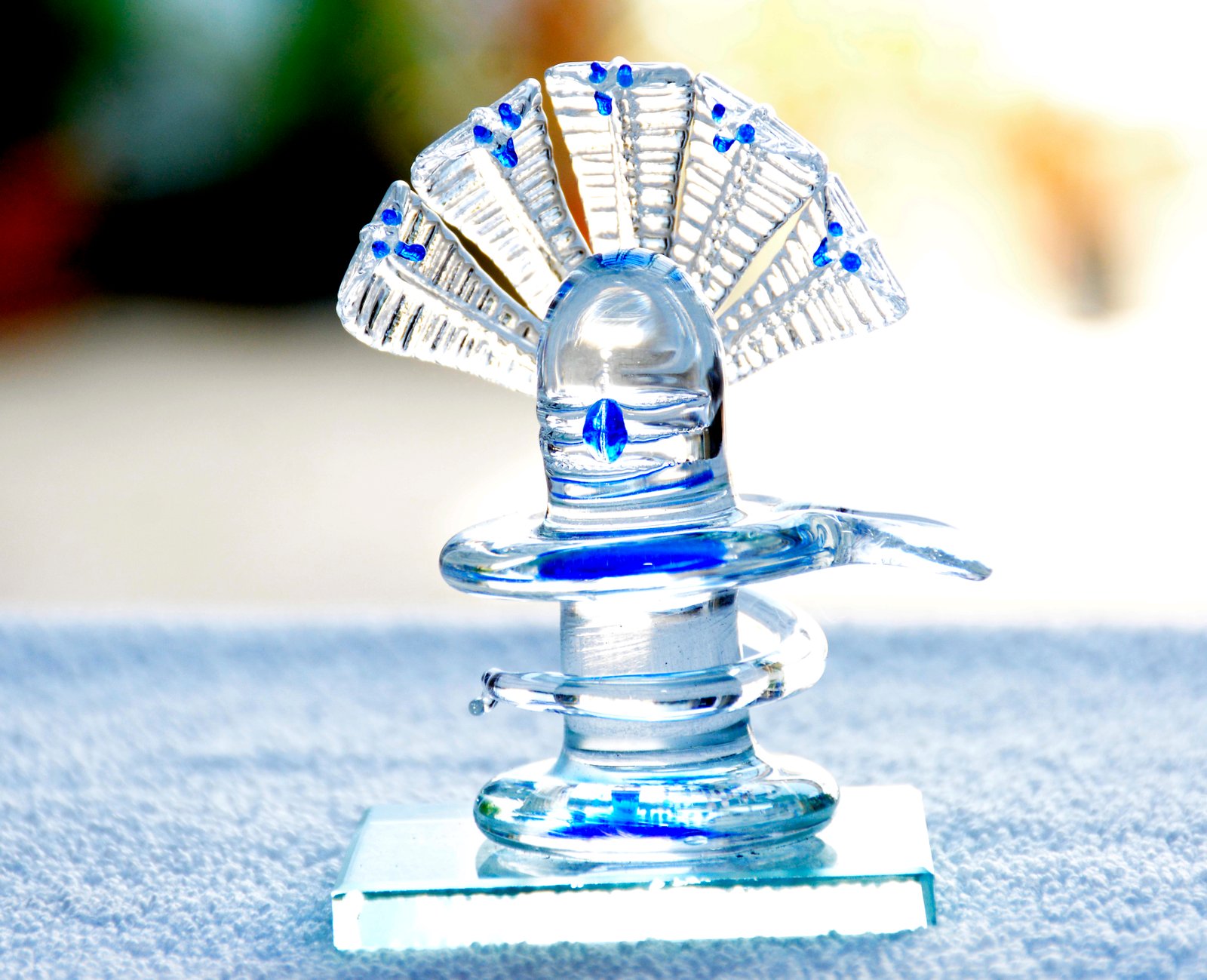 A small, decorative glass Shivling, featuring a combination of clear and blue glass elements, displayed on a glass base.